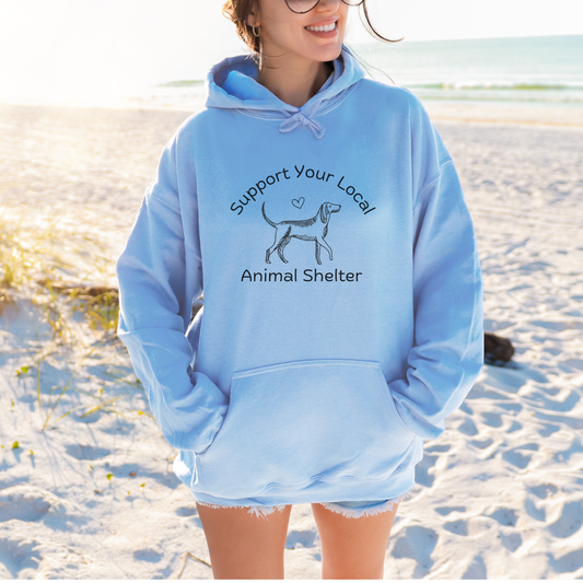 A woman standing on a sandy beach wearing a light blue hoodie with a black graphic that reads 'Support Your Local Animal Shelter' and shows a dog silhouette. The background shows the ocean and beach.