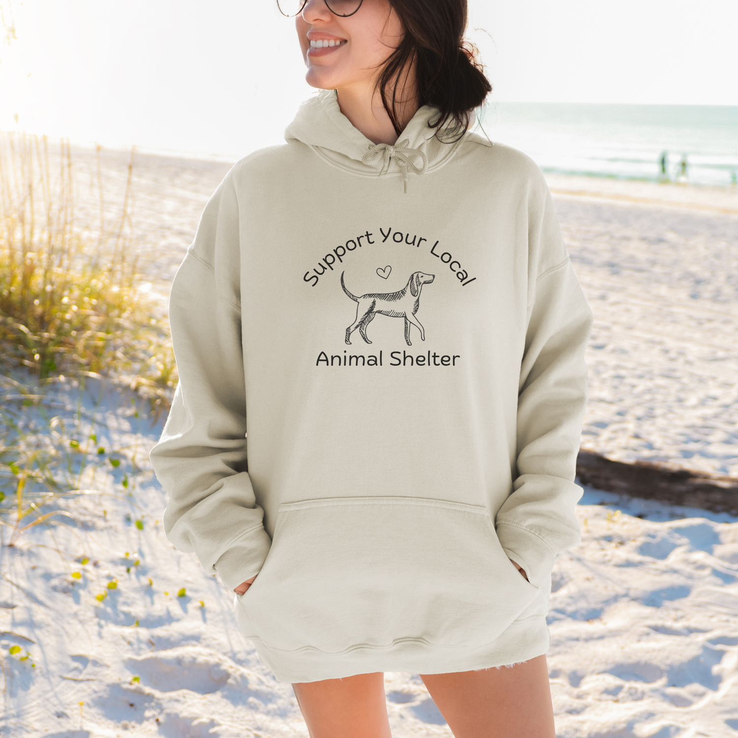 A woman wearing a light beige hoodie with a black graphic that reads 'Support Your Local Animal Shelter' stands on a sandy beach. The hoodie features a simple dog silhouette graphic in the center.