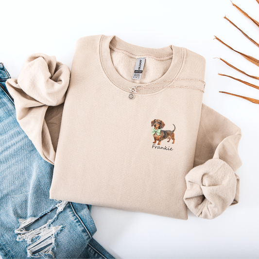 Beige sweatshirt with embroidered dog design and text, folded on a white surface with jeans.