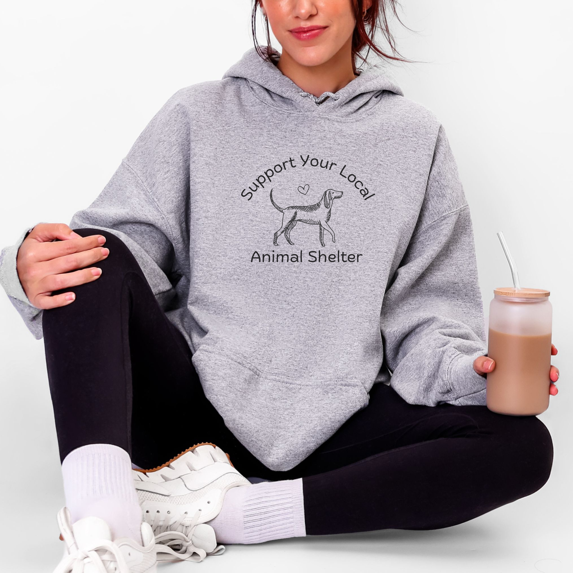 A woman sitting cross-legged wearing a heather gray hoodie with a black graphic that reads 'Support Your Local Animal Shelter' and shows a dog silhouette. She is wearing black leggings, white socks, and white sneakers, and holding a frosted tumbler with a brown drink against a plain white background.