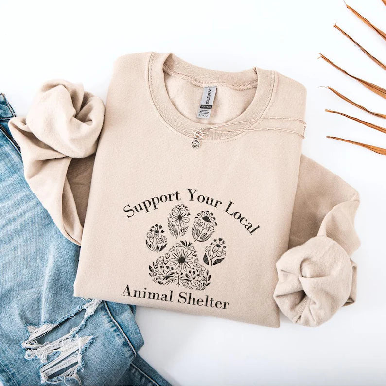 Beige sweatshirt with 'Support Your Local Animal Shelter' text and paw print design on a white background.